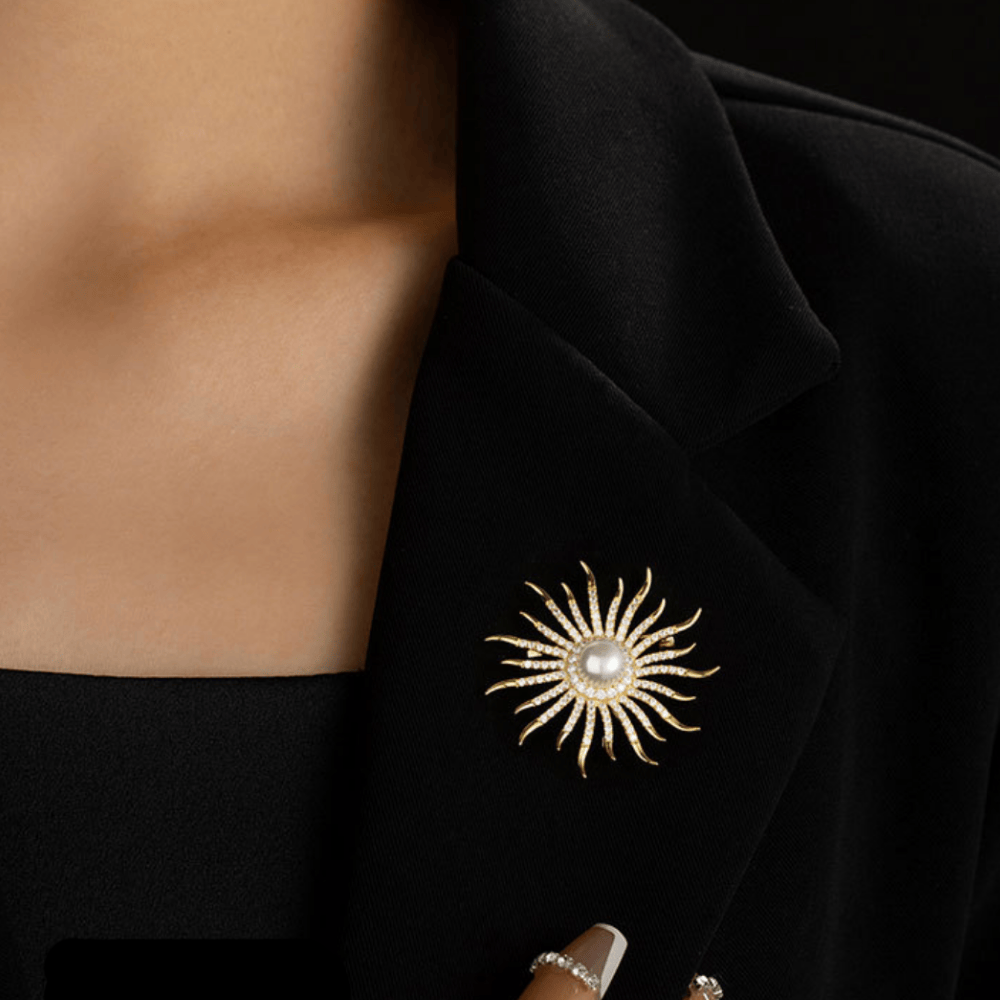 AVELIA gold sun brooch featuring a large central pearl surrounded by curved, crystal-lined rays on a black background.