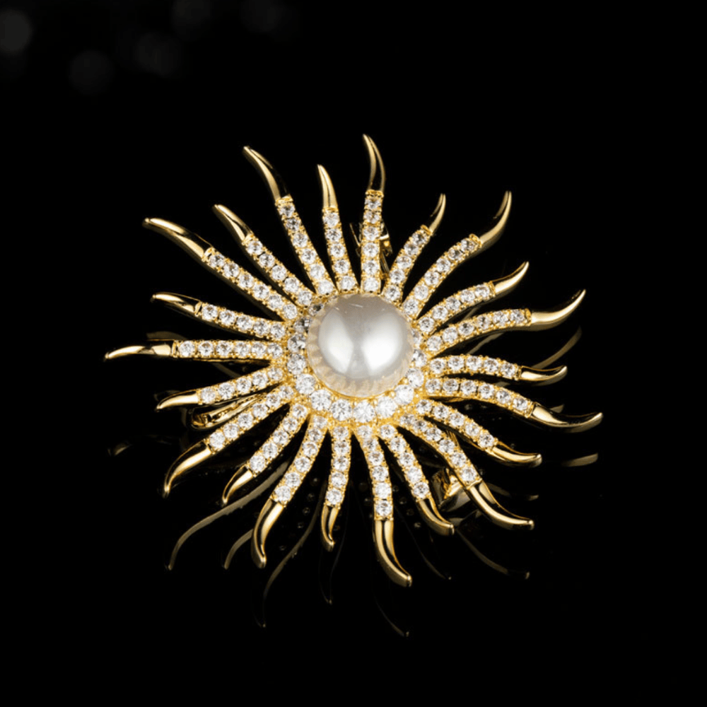 AVELIA gold sun brooch featuring a large central pearl surrounded by curved, crystal-lined rays on a black background.