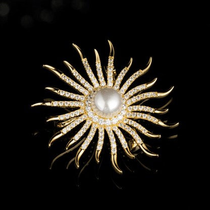 AVELIA gold sun brooch featuring a large central pearl surrounded by curved, crystal-lined rays on a black background.