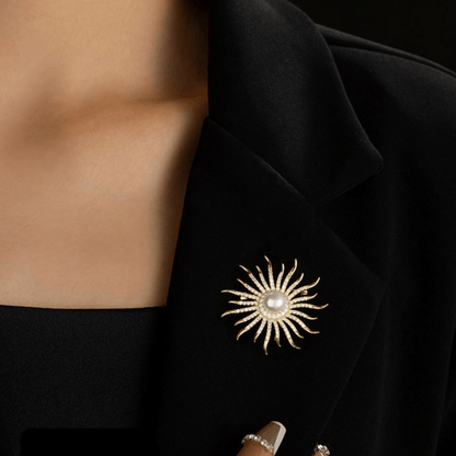 AVELIA gold sun brooch featuring a large central pearl surrounded by curved, crystal-lined rays on a black background.