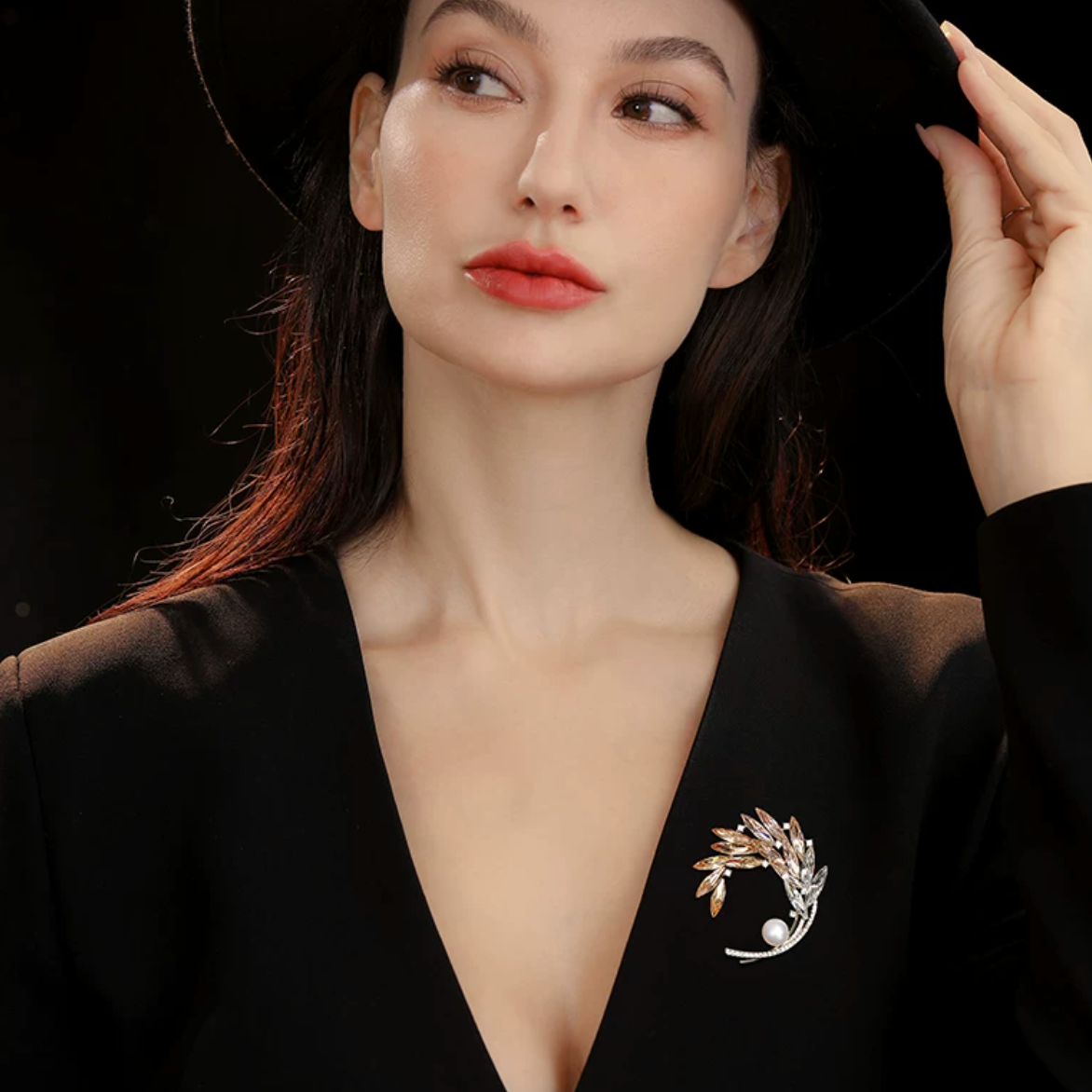 A woman wearing a black wide-brim hat and a black deep V-neck blazer, featuring a silver and gold leaf-shaped brooch with a pearl accent on the left chest, set against a solid black background.