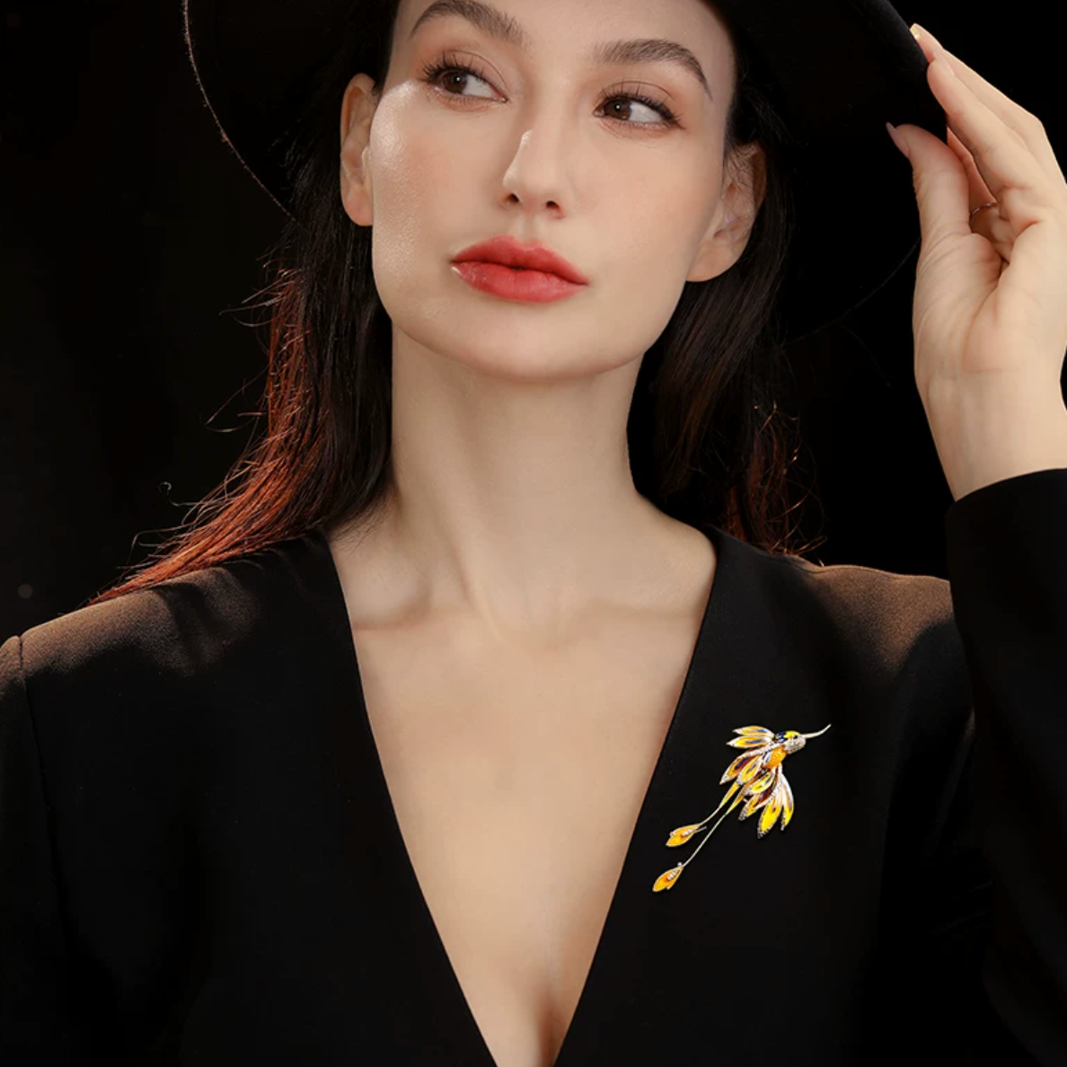 A woman wearing a black wide-brim hat and a black deep V-neck top, with a small gold and yellow floral brooch pinned to the chest. She is holding the brim of her hat against a solid black background.